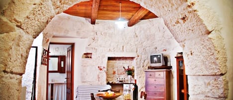 Una cocina rústica de piedra con techo de madera, una mesa de comedor cubierta con mantel escocés y un televisor sobre un mueble de madera.