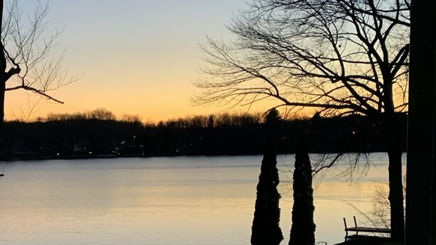 A serene lakeside view with a clear sky, calm water, and a grassy area with a wooden fence. - Gorgeous newer home on Newton Lake/Crystal Lake near Elk mountain ski resort.  (Greenfield Township)