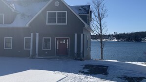 A two-story house with a red door and white-framed windows, surrounded by snow-covered ground and a clear sky. - Gorgeous newer home on Newton Lake/Crystal Lake near Elk mountain ski resort.  (Greenfield Township)