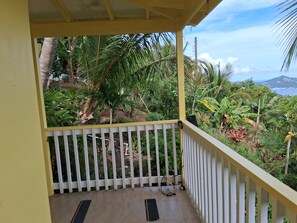 A balcony with a white railing, a tiled floor, and a view of lush greenery and a distant mountain.