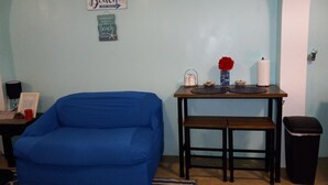 A room with a blue sofa, a small table with decorative items, a trash bin, and wall decorations. - Beautiful Apartment w- gorgeous expansive views, beautiful fruit trees & gardens (ST. THOMAS)