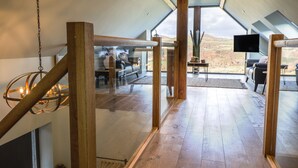 A spacious loft-style room with a high ceiling, wooden beams, and a large window offering a view of the countryside. - Tyn Llwyn Barn (Corwen)