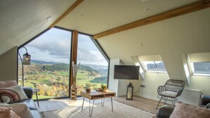 A spacious loft bedroom with a large skylight, a flat-screen TV, and a view of the countryside. - Tyn Llwyn Barn (Corwen)