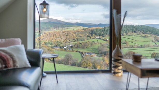 A room with a large window overlooking a valley, a sofa, a small table, and a decorative vase. - Tyn Llwyn Barn (Corwen)