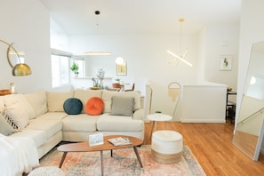 Uma sala de estar moderna com um sofá seccional, uma mesa de centro e uma pequena mesa lateral. Há uma escada que leva a um andar superior, e uma área de jantar com mesa e cadeiras.