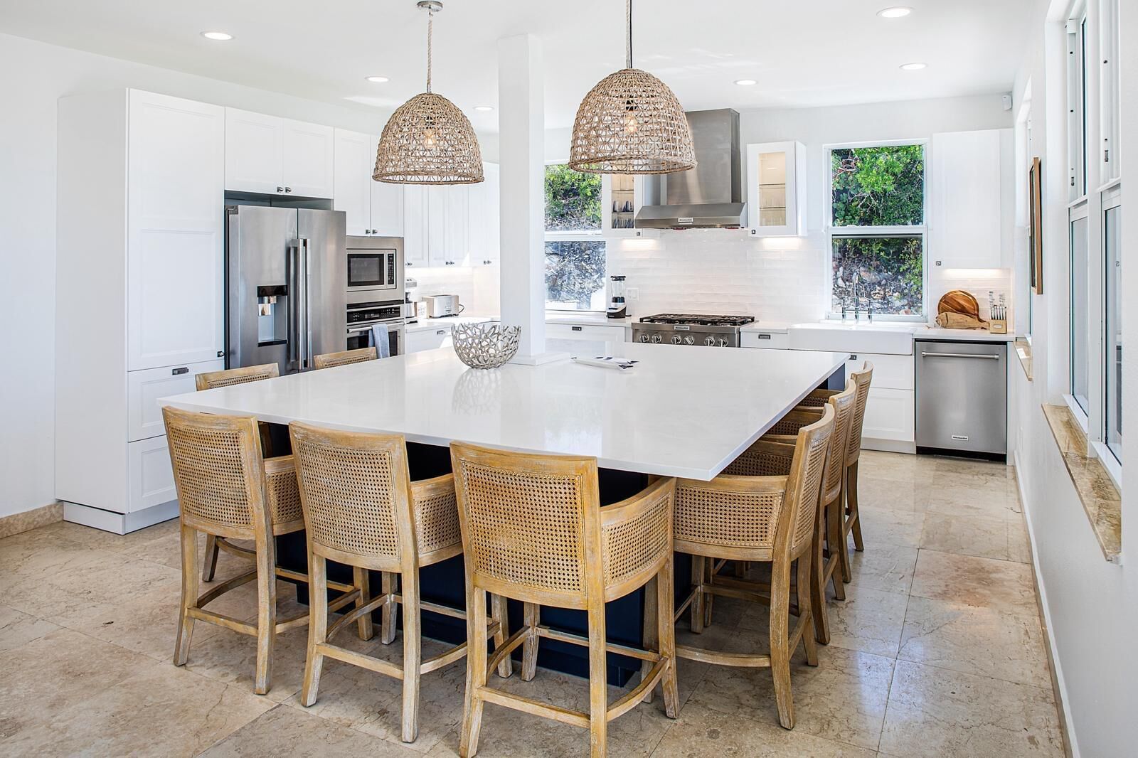 A modern kitchen with white cabinetry, a large island, wicker chairs, and pendant lighting.
