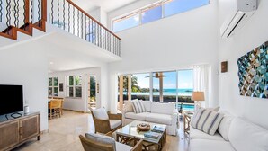 Ein geräumiges Wohnzimmer mit großem Fenster mit Blick auf einen Strand, ausgestattet mit einer Couch, Sesseln und einem Couchtisch.
