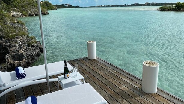 Eine Holzterrasse mit weißen Liegestühlen und einem Tisch, mit Blick auf klares türkisblaues Wasser.