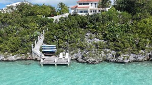 Eine Holzsteg mit Liegestühlen und einem Boot, der zu einem Haus inmitten üppiger Vegetation und klarem türkisblauem Wasser führt.