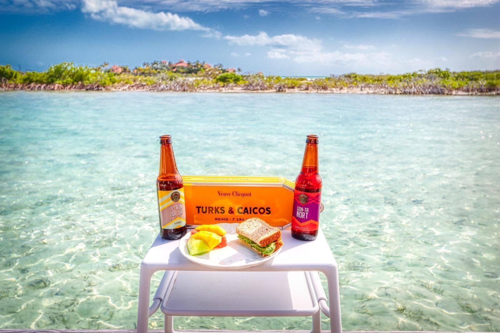 Una mesa con una caja de turcs i caicòs, dos botellas de cerveza y un plato con comida y una bebida en la playa.