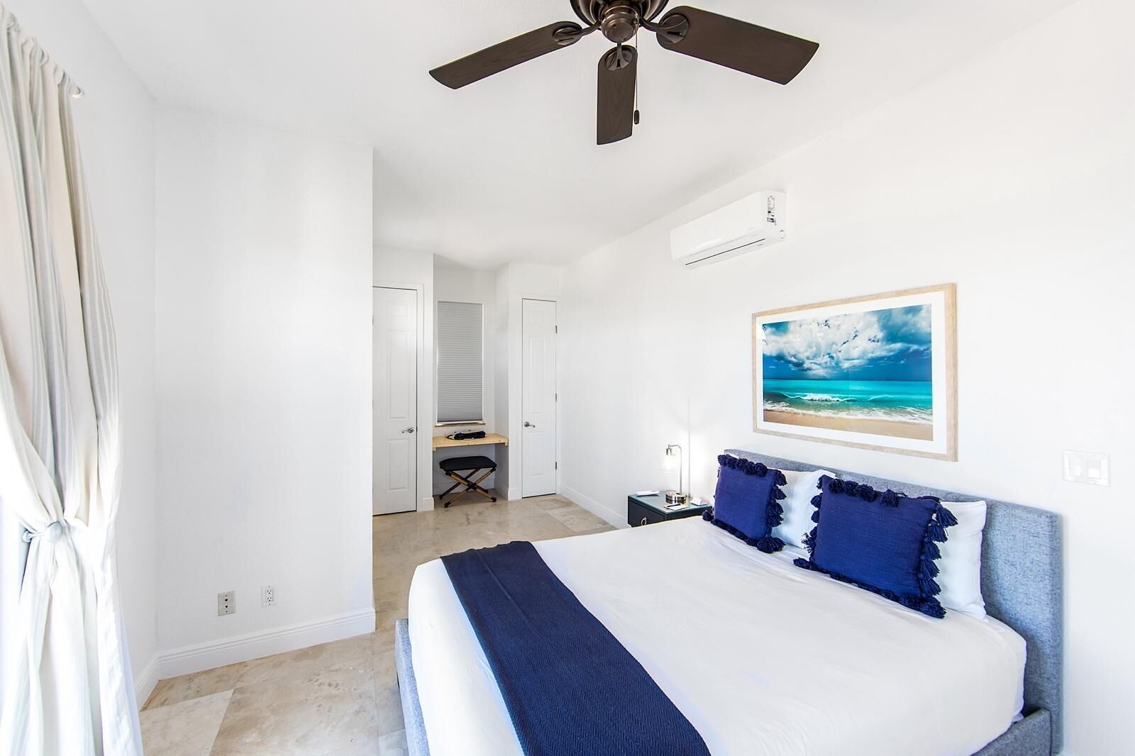 A bedroom with a bed, a ceiling fan, an air conditioning unit, a desk with a chair, and a framed picture on the wall.