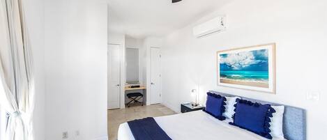 A bedroom with a bed, a ceiling fan, an air conditioning unit, a desk with a chair, and a framed picture on the wall.