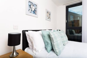 A neatly made bed with white and teal pillows, a black lamp on a wooden nightstand, and two framed artworks on the wall.