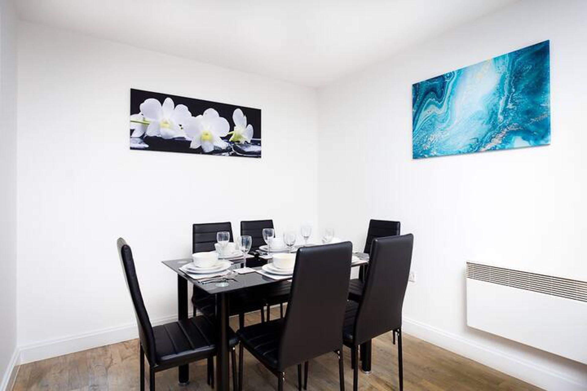 Een eetgedeelte met een tafel voor vier personen, zwarte stoelen en een witte muur met twee kunstwerken in lijsten.