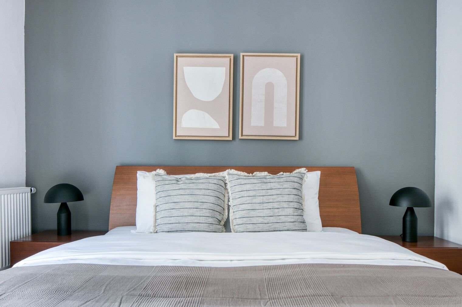Un lit bien fait, avec une tête de lit en bois, deux oreillers et une couverture grise. Il y a deux tables de chevet avec des lampes noires. Le mur est peint en gris, et deux œuvres d’art abstraites encadrées sont accrochées au-dessus du lit.
