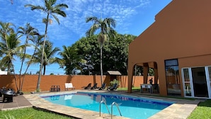 A swimming pool with lounge chairs and a table, surrounded by palm trees and a building with large windows. - Ponta dos Corais (Guarujá)