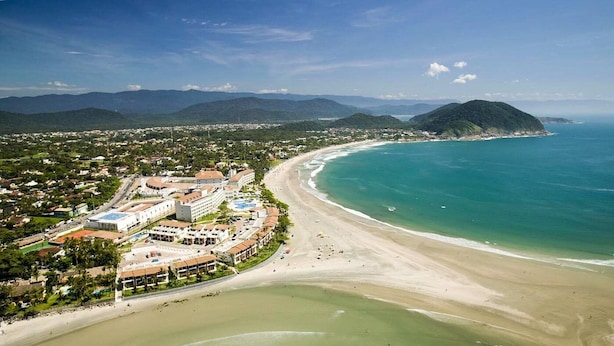 An aerial view of a coastal resort with a beachfront, a large hotel complex, and surrounding greenery. - Ponta dos Corais (Guarujá)