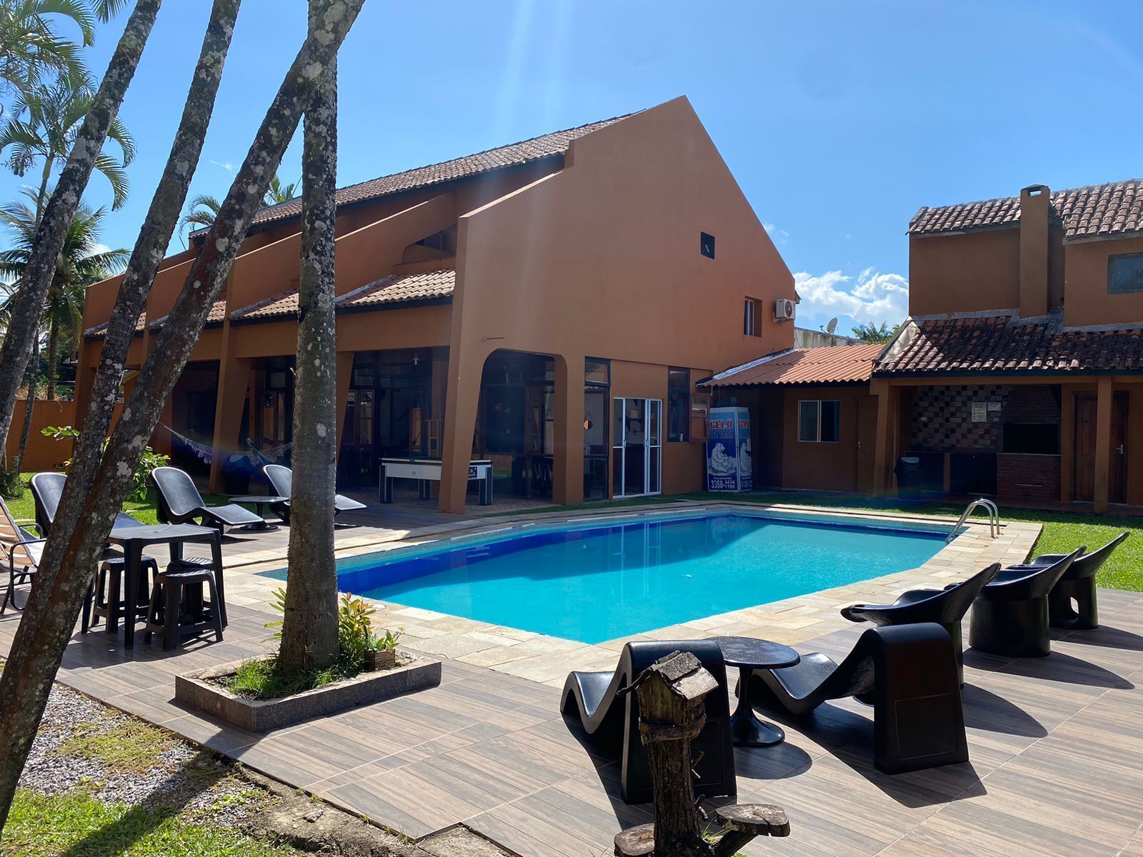 Un área de piscina con sillones de descanso y una mesa, rodeada de palmeras y un edificio con techo de tejas.