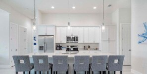A modern kitchen with white cabinets, a stainless steel refrigerator, a microwave, and a built-in oven. There are four grey upholstered chairs around a central island with a sink.