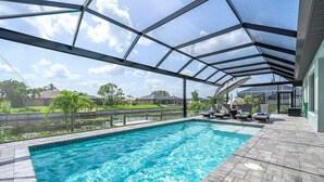 Una piscina con cerramiento de vidrio, rodeada por un patio con sillas de descanso y vistas a un barrio residencial.