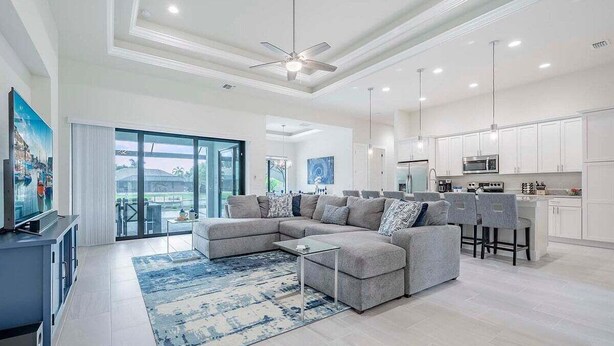 Una sala de estar moderna con un sofá seccional, una mesa de centro y un televisor. Hay una zona de cocina con gabinetes blancos y una barra.