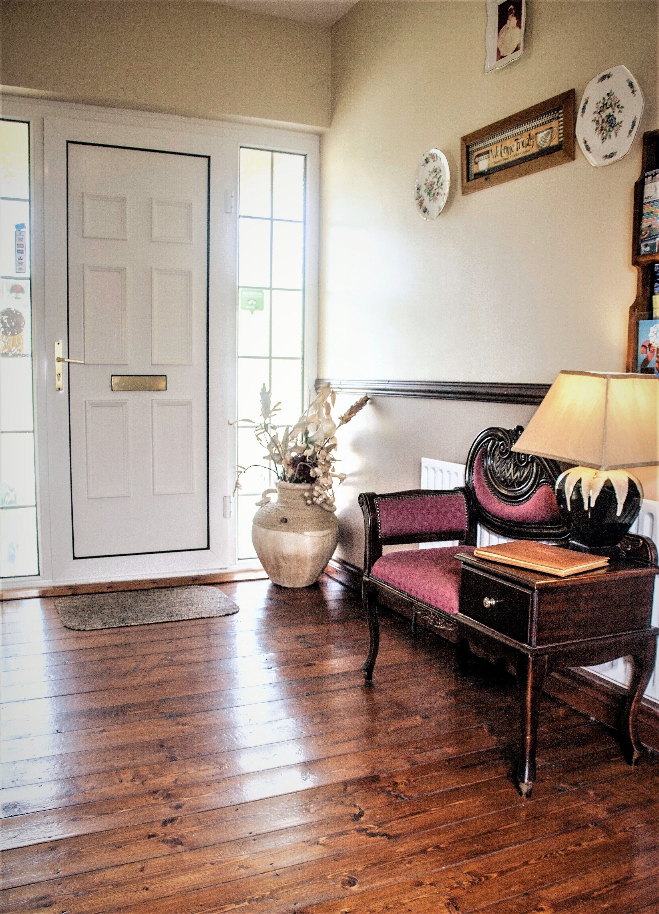 A room with a wooden floor, a white door, a vase with dried flowers, a purple upholstered chair, a wooden nightstand, and a lamp.