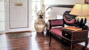A room with a wooden floor, a white door, a vase with dried flowers, a purple upholstered chair, a wooden nightstand, and a lamp.