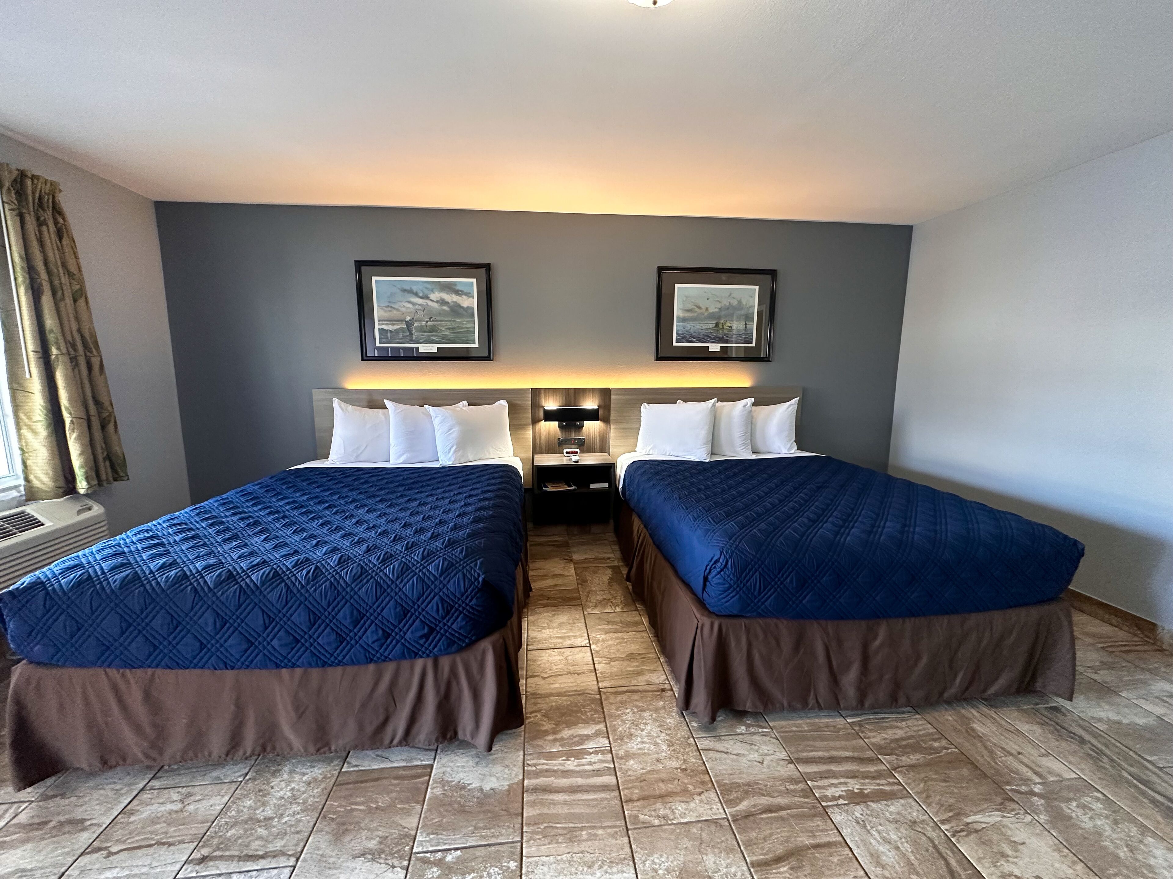 A hotel room with two beds, a nightstand, a lamp, and two framed pictures on the wall.