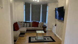 Een woonkamer met een hoekbank, een bijzettafeltje en een flatscreen televisie aan de muur.