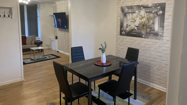 Een eetruimte met een donker houten tafel, zwarte stoelen en een witte vaas met groen. Een flatscreen televisie is aan de muur bevestigd, en daarboven hangt een lijst met een bloemenafbeelding.