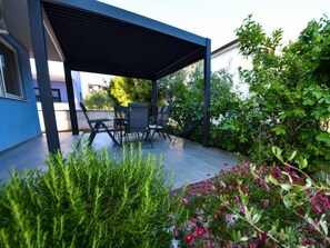 Ein überdachter Patio-Bereich mit Gartenmöbeln, umgeben von üppiger Bepflanzung und blühenden Pflanzen.
