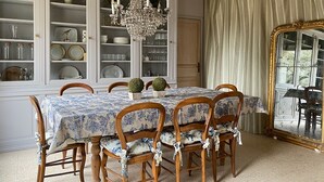 A dining room with a table set for six, surrounded by wooden chairs with floral upholstery. A large white cabinet with glass doors displays various dishes and glassware. A chandelier hangs from the ceiling, and a large mirror with a gold frame is on the wall. - Stylish farmhouse with pool, sleeps 8-10 (Verneuil-sur-Indre)