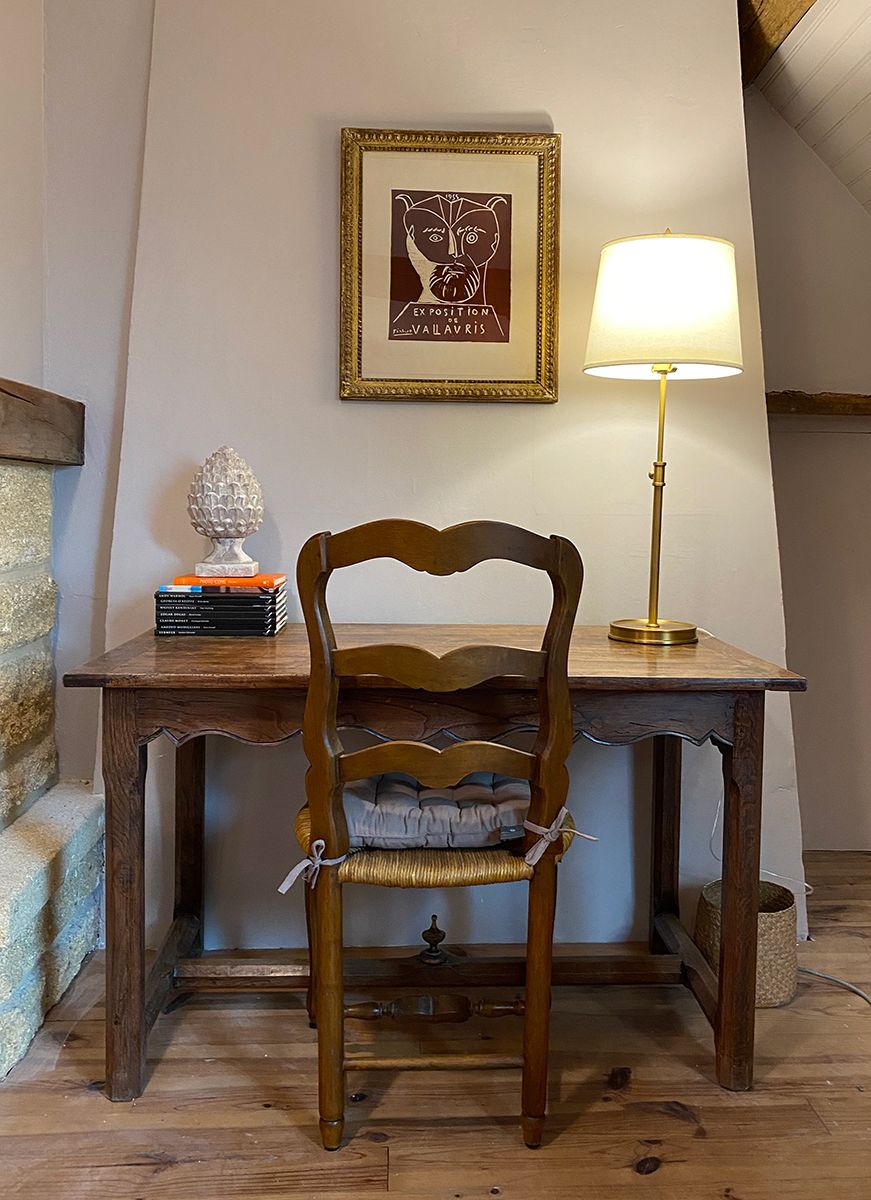 Una silla de madera con asiento y respaldo acolchados, ubicada frente a un escritorio con una lámpara y un cuadro en la pared.