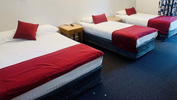 Een hotelkamer met drie bedden, elk voorzien van witte beddengoed en rode accenten. Daartussen staat een houten nachtkastje met een telefoon en een lamp.