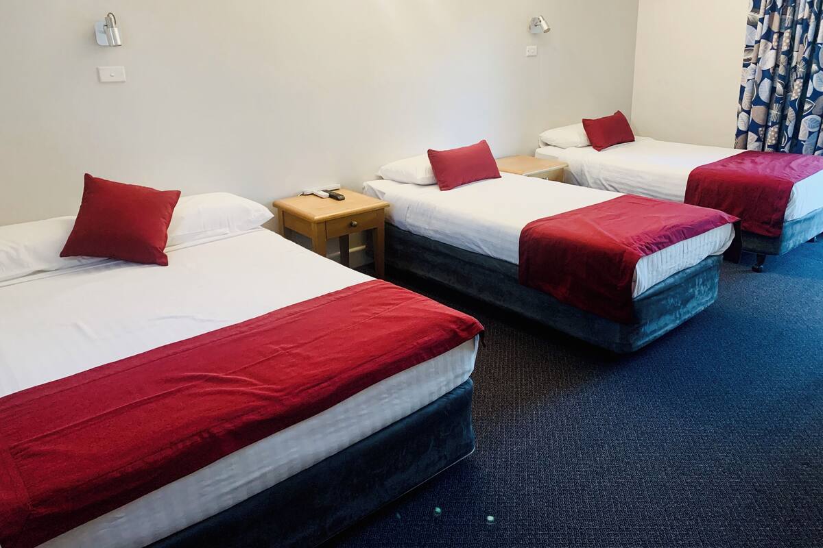 Une chambre d’hôtel avec trois lits, chacun agrémenté de draps blancs et de touches de couleur rouge. Une table de chevet en bois, sur laquelle se trouvent un téléphone et une lampe, est placée entre les lits.