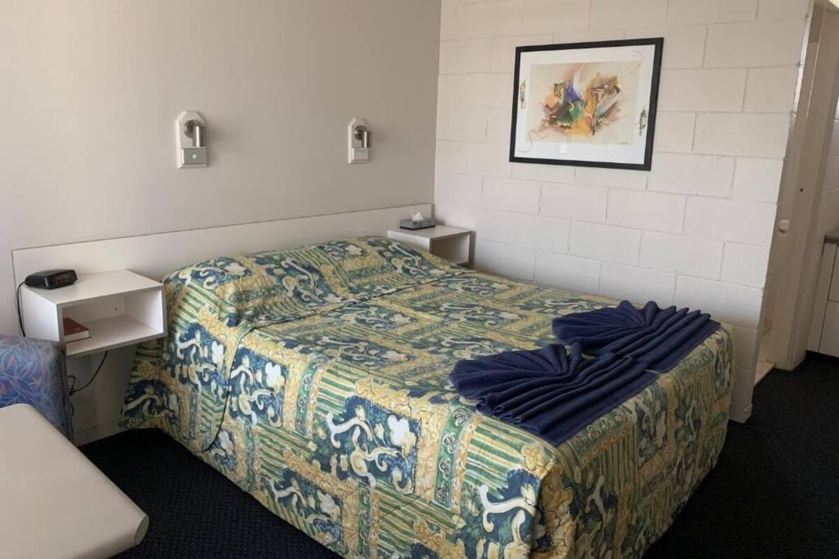 Une chambre d’hôtel avec un lit, un bureau et une chaise. Il y a un tableau encadré au mur et une porte sur la droite.