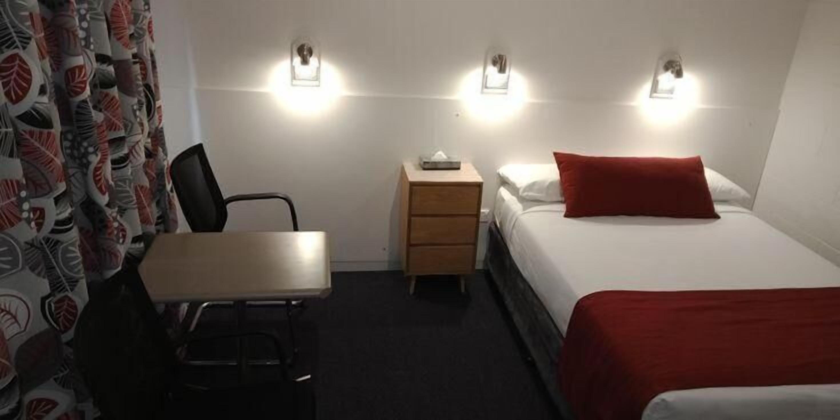 Une chambre d’hôtel comprenant un lit, un bureau, une chaise, une table de chevet et un luminaire fixé au mur.