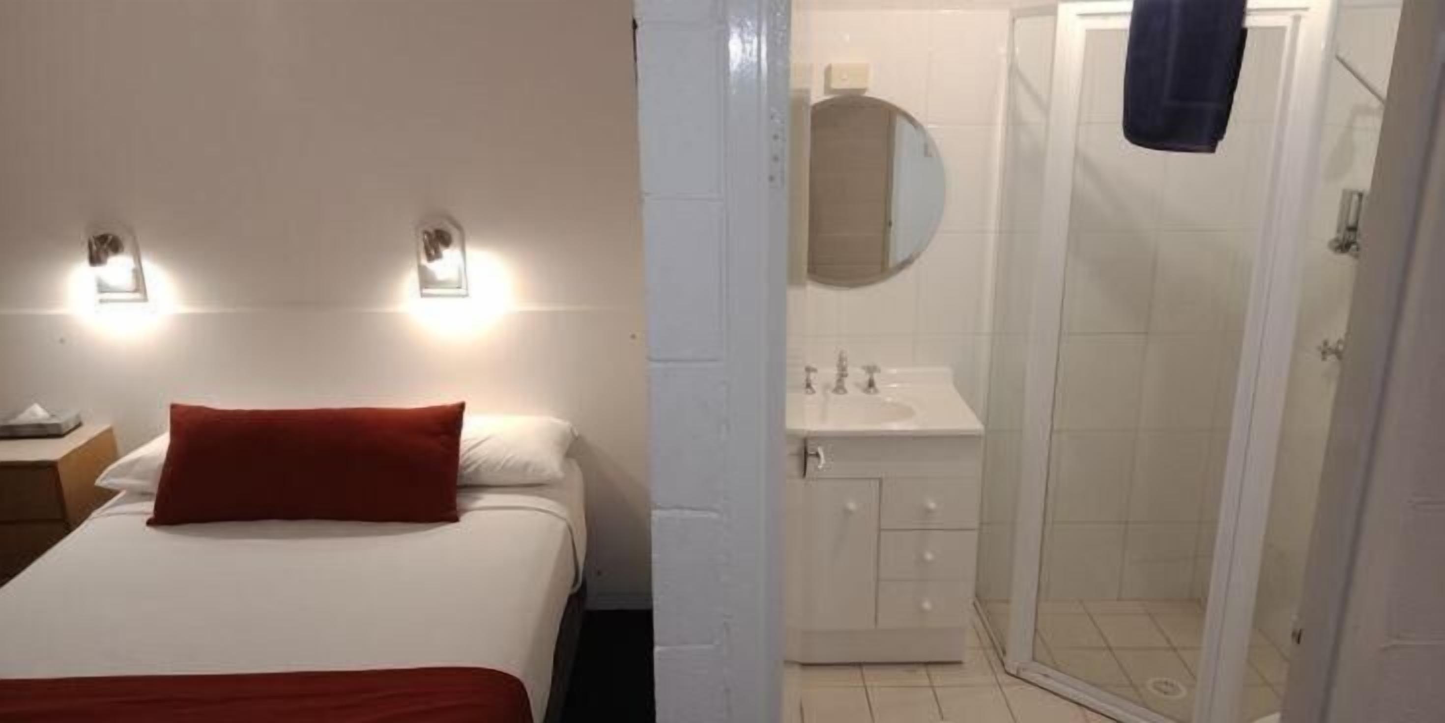 A hotel room with a bed, a nightstand, a wall-mounted light fixture, a bathroom with a sink, mirror, and shower.