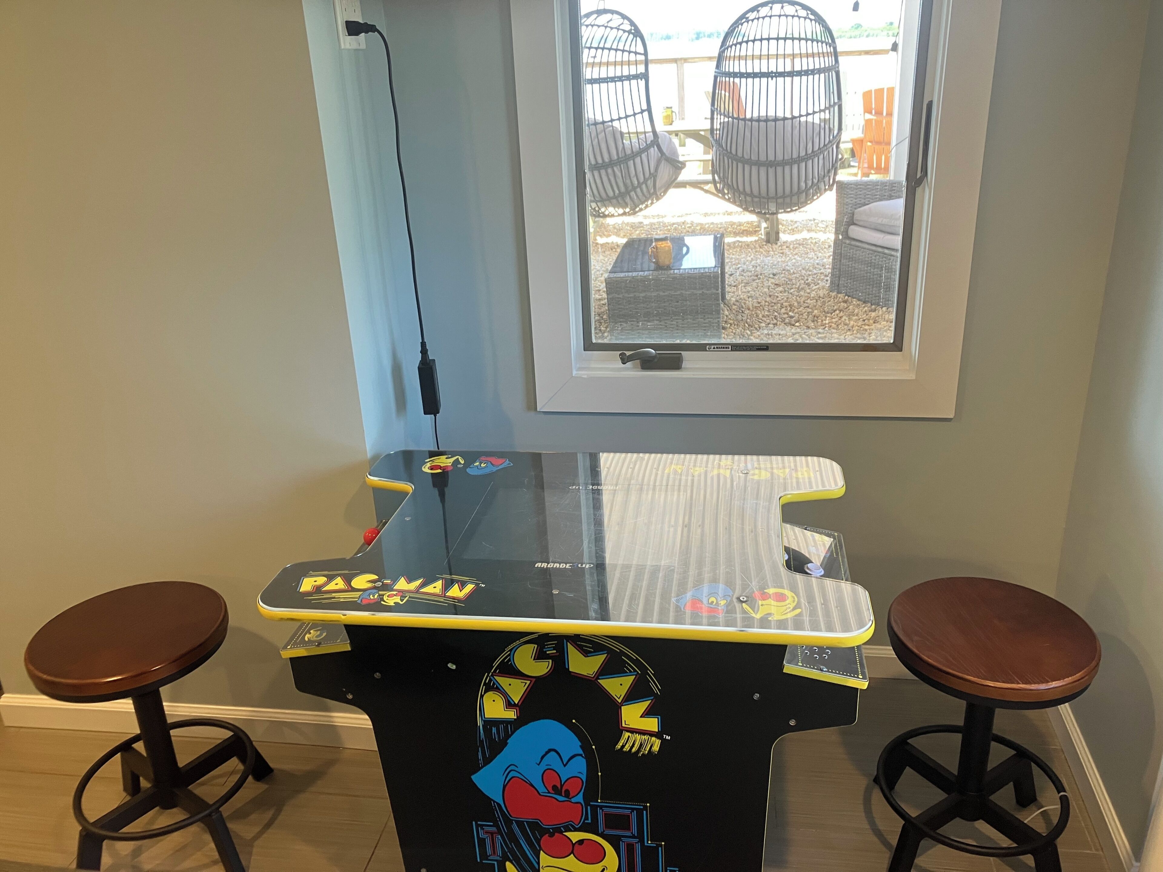 Una máquina arcade de Pac-Man con dos taburetes, una ventana que da a una terraza al aire libre, y un cable de alimentación conectado.