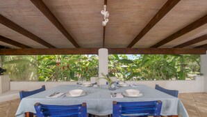 A covered outdoor dining area with a table set for four, surrounded by chairs, and a view of lush greenery through a glass wall. - Villa TORU beautiful tropical style villa with outdoor bath, on the sunset side (Rarotonga)