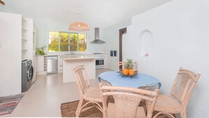 A modern kitchen with white cabinetry, a central island, and a dining area with wicker chairs and a blue tablecloth. - Villa TORU beautiful tropical style villa with outdoor bath, on the sunset side (Rarotonga)