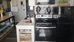 Cocina compacta con microondas, horno y refrigerador negros. Hay un lavamanos con grifo, un estante de madera con utensilios de cocina y una pequeña ventana.