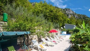 A sunlit outdoor seating area with lounge chairs and a covered bench, surrounded by lush greenery and a mountain backdrop. - Vacation home Karin by Interhome (Lovran)
