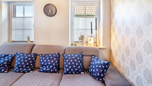 A living room with a sofa, patterned pillows, a wall clock, and a window with blinds. - Vacation Home 'Segelboot' with Garden, BBQ & Wi-Fi (Krummhörn)