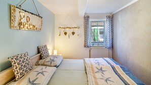 A bedroom with two beds, a decorative wall hanging, a window with patterned curtains, and a hanging ornament. - Vacation Home 'Segelboot' with Garden, BBQ & Wi-Fi (Krummhörn)