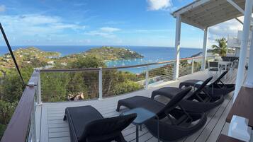 A covered outdoor seating area with wicker furniture and a view of the sea and a hilly landscape.