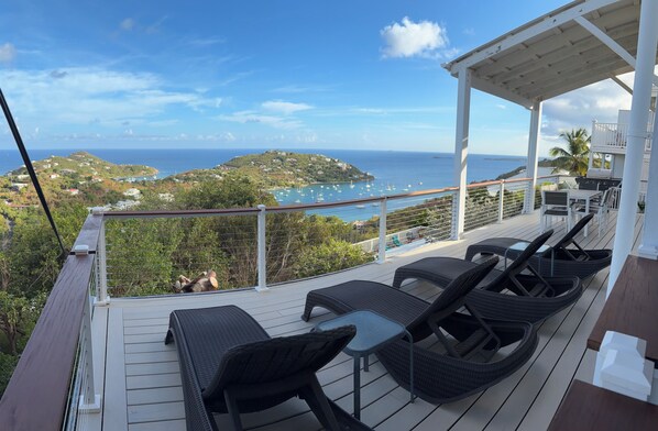 A covered outdoor seating area with wicker furniture and a view of the sea and a hilly landscape.