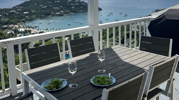 Une terrasse en bois avec une table dressée pour deux personnes, offrant une vue sur la côte, avec des bateaux et une île au loin.