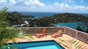 Un balcon avec une piscine, deux chaises longues et une vue sur un paysage côtier, avec des maisons et la mer.