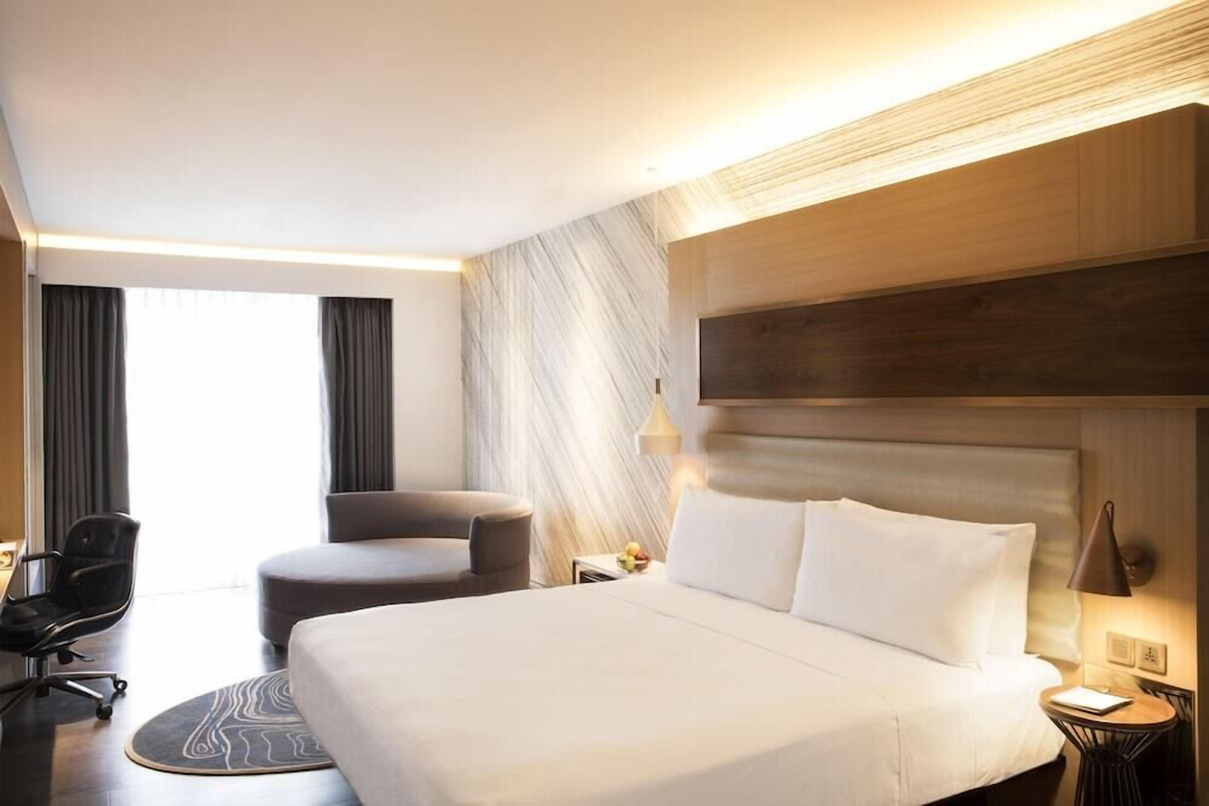 A modern hotel room with a large bed, a desk, a chair, and a small table.